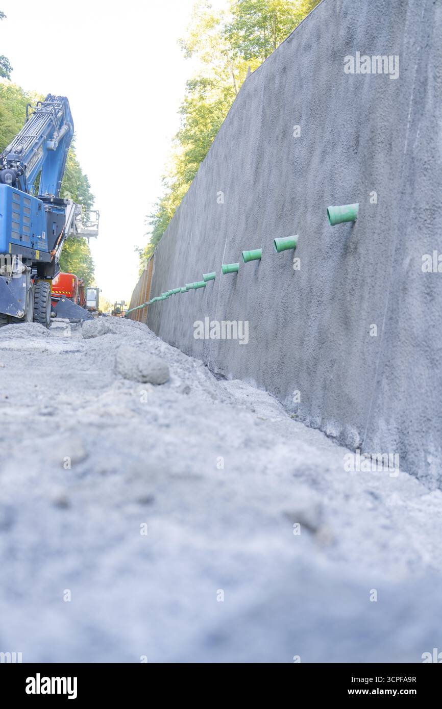 Grande parete in cemento e escavatore blu su una strada di ghiaia, cantiere Hermann Hessebahn, Calw, Germania Foto Stock