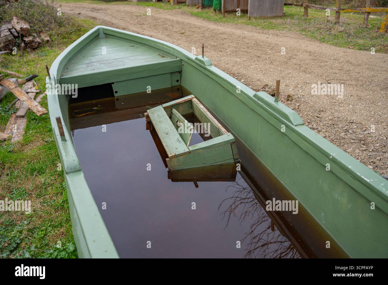 Vecchio peschereccio verde con timone arrugginito vicino al sentiero rurale. Barca di legno verde inondata piena di acqua, sommersa. Campagna Foto Stock