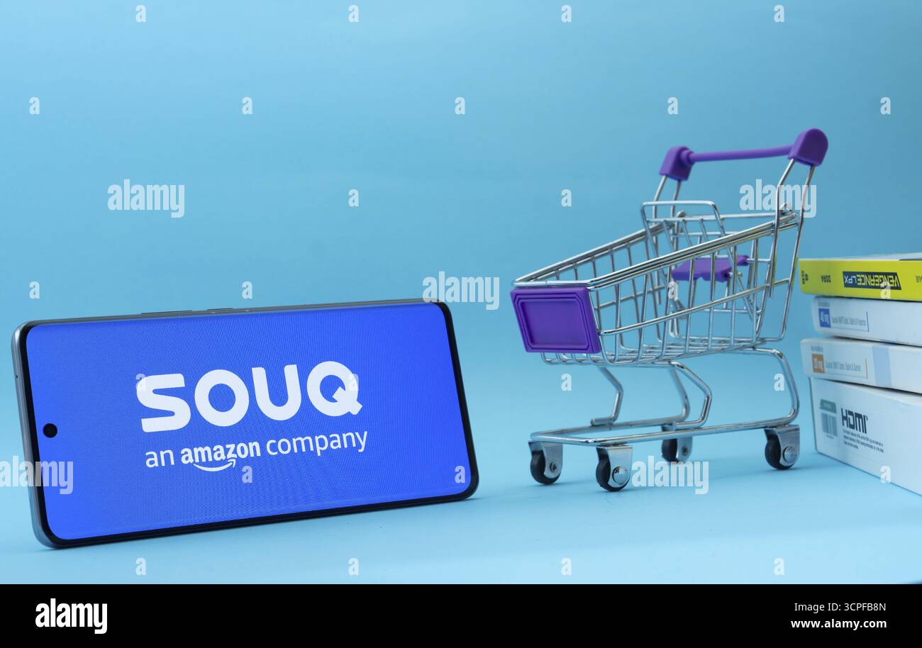 Dhaka, Bangladesh - 3 luglio 2025: Smartphone con logo Souq accanto a un piccolo carrello e scatole su sfondo blu Foto Stock