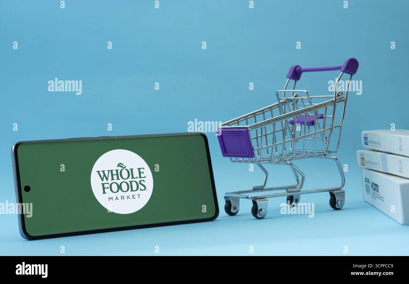 Dhaka, Bangladesh - 3 luglio 2025: Smartphone con logo Whole Foods Market accanto a un piccolo carrello e scatole su sfondo blu Foto Stock