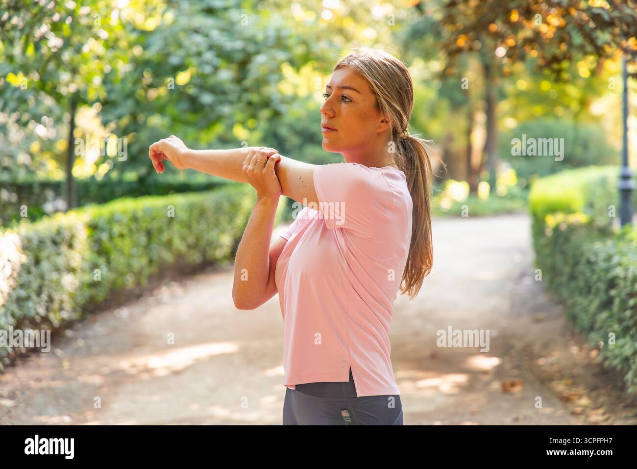 Giovane donna bionda che esegue un braccio pre-esercizio si allunga all'aperto su un percorso del parco, si prepara per il suo allenamento e mantiene una sana rotta di fitness Foto Stock
