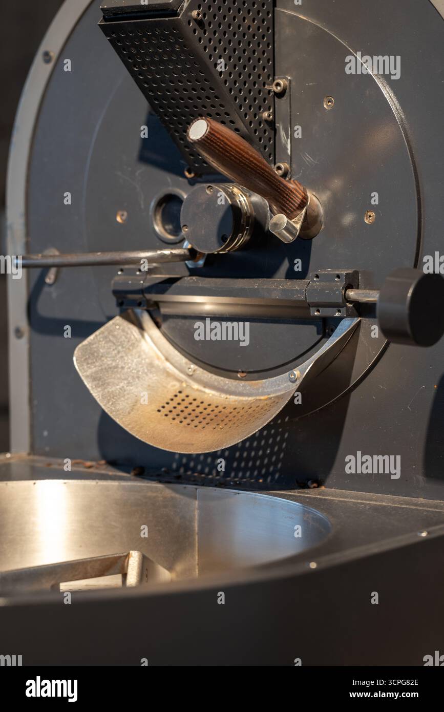 Fotografia in primo piano verticale di una torrefazione con tamburo metallico, impugnatura e scivolo. La macchina si trova in un ambiente di officina, pronta per il roa Foto Stock