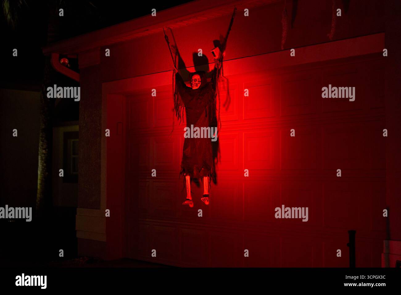 Un mietitore con cappuccio e gambe ossee è appeso alla porta di un garage sotto una luce rossa profonda. Spettrale scenografia da cortile di Halloween con ombre spettacolari di notte. Foto Stock