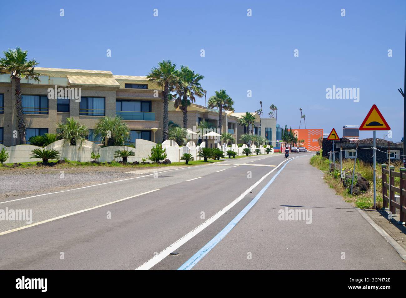 Le moderne ville e le palme costeggiano una strada sul mare segnata dalla pista ciclabile blu di Jeju, mentre pareti basse, giardini e cartelli di avvertimento seguono il lava Coas Foto Stock