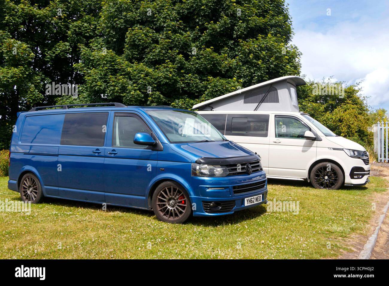 Due camper volkswagen, un T5 blu e un T6 grigio, parcheggiati su un'area erbosa con alberi sullo sfondo durante una giornata di sole Foto Stock