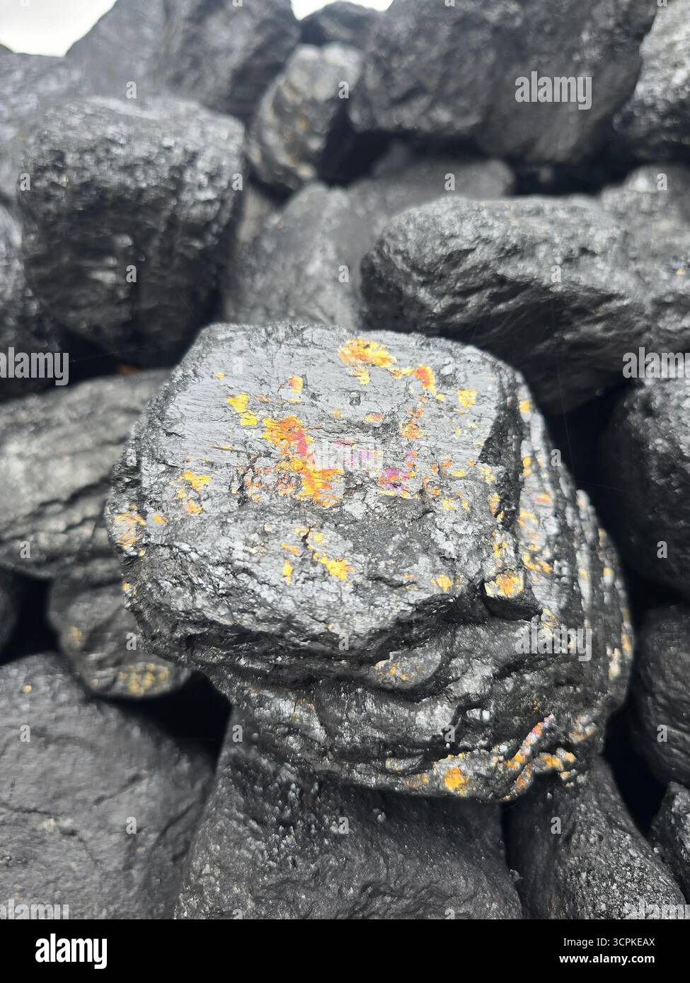 Primo piano di una grande pila di grumi di carbone nero lucido, con un pezzo che mostra in modo prominente strisce colorate, iridescenti e simili a minerali. - Immagine stock catturata con smartphone