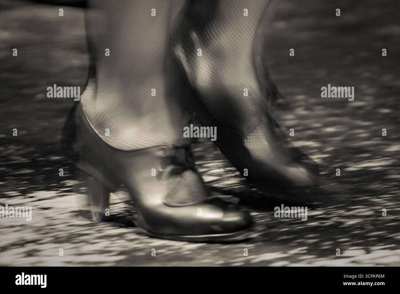 Un primo piano, sfocato, in bianco e nero mostra le gambe e i piedi di una persona in calze a rete nere e scarpe con tacco nero, muovendosi rapidamente su un pavimento a motivi geometrici, magari ballando. Foto Stock