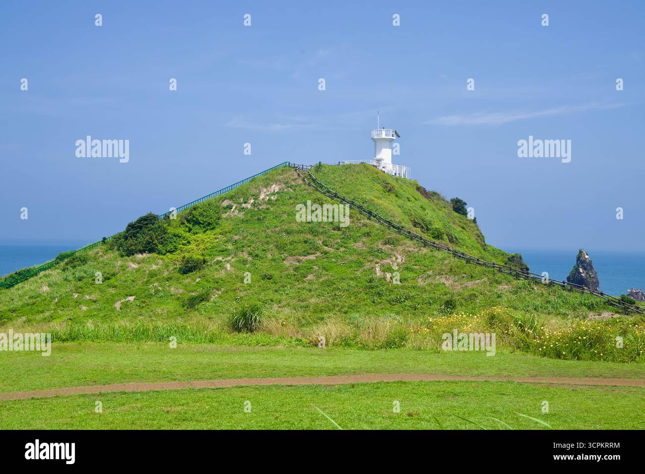Un faro bianco incornicia la collina erbosa di Seopjikoji, raggiunta da scalinate di legno che salgono la cresta sopra le scogliere marine e i faraglioni al largo. Foto Stock