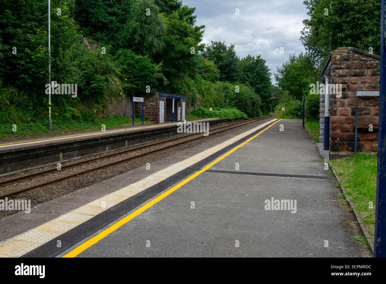 Vista tranquilla della stazione ferroviaria di Ince ed Elton, Cheshire, con piattaforme, binari e ripari originali in pietra circondati da vegetazione lussureggiante. Foto Stock