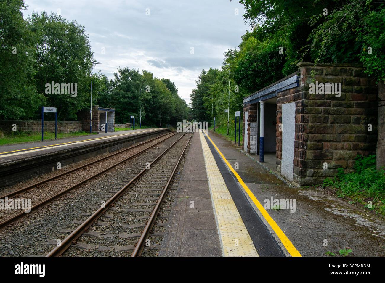 Vista tranquilla della stazione ferroviaria di Ince ed Elton, Cheshire, con piattaforme, binari e ripari originali in pietra circondati da vegetazione lussureggiante. Foto Stock