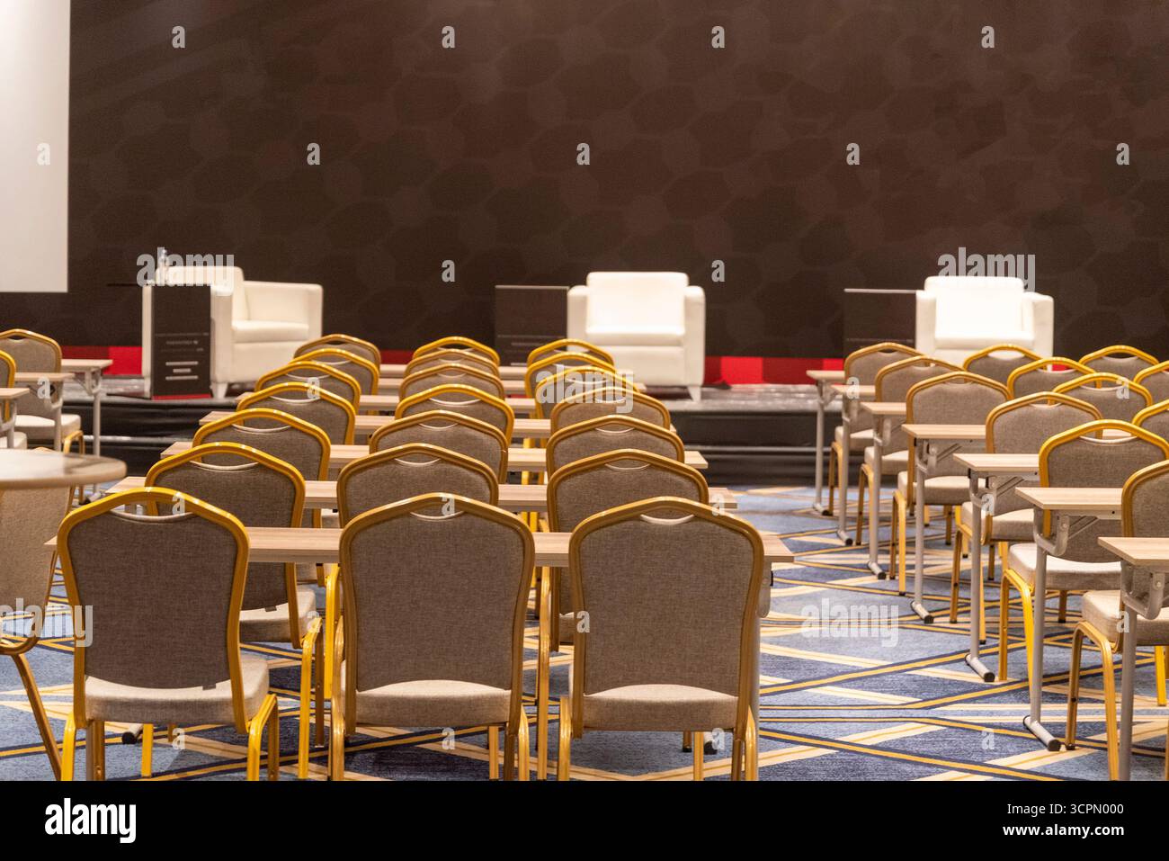 Le sedie e i tavoli sono ordinatamente disposti in fila, preparati per una conferenza imminente. Due poltrone bianche si trovano ben visibili sul palco, invitanti altoparlanti Foto Stock