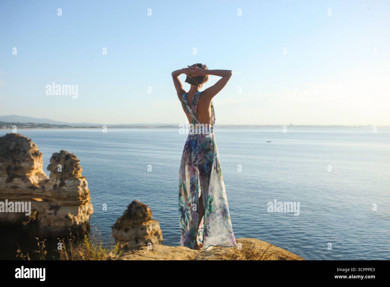 Donna irriconoscibile con un lungo abito floreale estivo in piedi davanti alla macchina fotografica con le braccia sollevate toccando i capelli e guardando splendide formazioni rocciose. Foto Stock