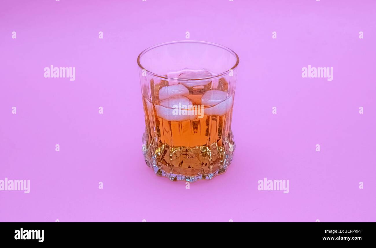 Elegante bicchiere riempito con una vivace bevanda color ambra e cubetti di ghiaccio su sfondo rosa brillante. Perfetto per i temi legati ai cocktail e agli eventi estivi. Foto Stock