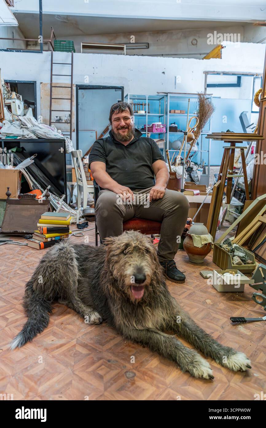 Uomo barbuto seduto in uno studio d'arte disordinato con il suo grande cane che riposa sul pavimento Foto Stock