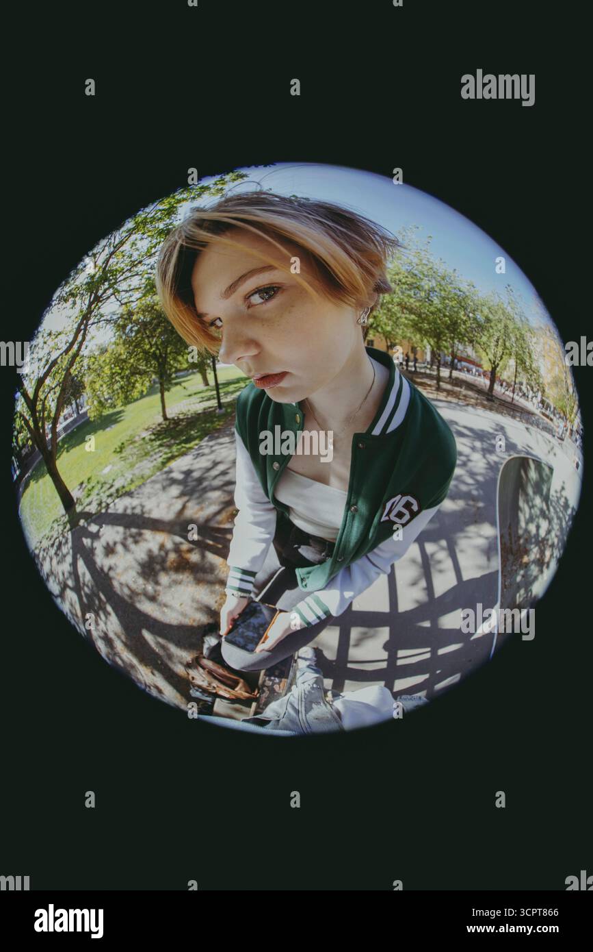 Ritratto ad angolo alto di un giovane con un'espressione vuota che tiene lo smartphone seduto su una ringhiera nella foto Fish Eye Foto Stock