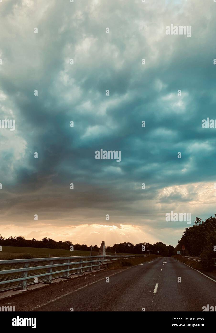 Strada rurale sotto le spettacolari nuvole di tempesta con orizzonte luminoso e spalle alberate - Immagine stock catturata con smartphone
