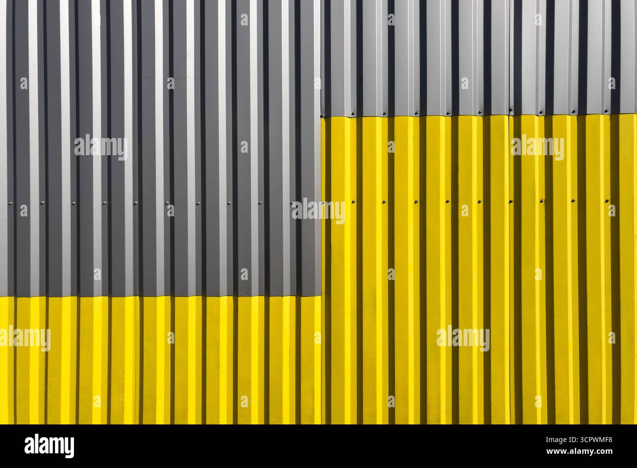 Parete ondulata industriale con linee verticali, metà verniciata in grigio e metà in giallo brillante, creando un forte contrasto geometrico simmetrico Foto Stock