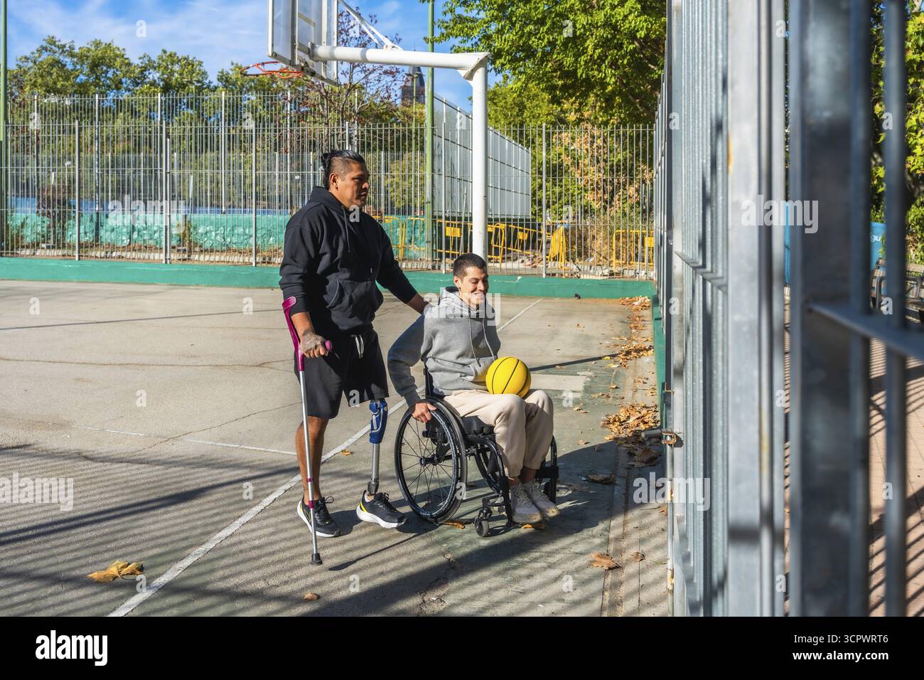 Amici che festeggiano l'inclusione sportiva su un campo da basket all'aperto, un uomo con stampelle e una protesi alle gambe si regge mentre spinge il sorriso Foto Stock