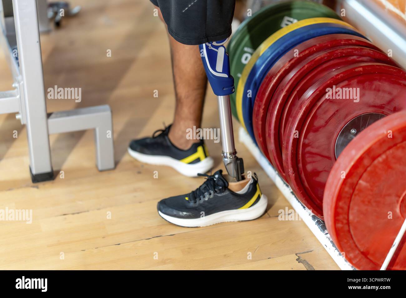 Uomo amputato in piedi su un pavimento di legno in una palestra, con una moderna gamba protesica e una normale scarpa sull'altro piede, accanto a una cremagliera di peso colorato Foto Stock