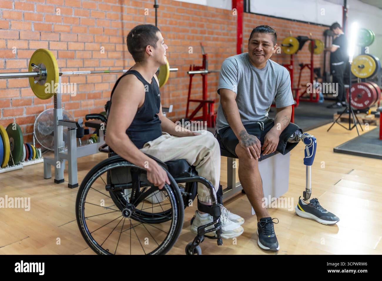 Due uomini disabili, uno su sedia a rotelle e l'altro con una protesi gamba, sorridono mentre si rilassano e parlano in palestra, evidenziando l'adattabilità fisica Foto Stock