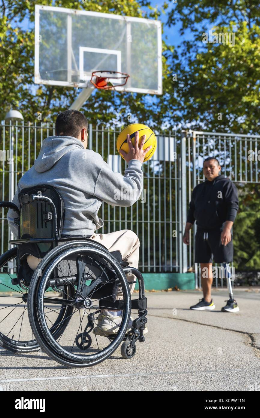 Uomo in sedia a rotelle che mira a sparare a un pallacanestro mentre il suo amico maschio, che usa una gamba protesica, sta a guardare su un campo da basket all'aperto, rappresentante Foto Stock