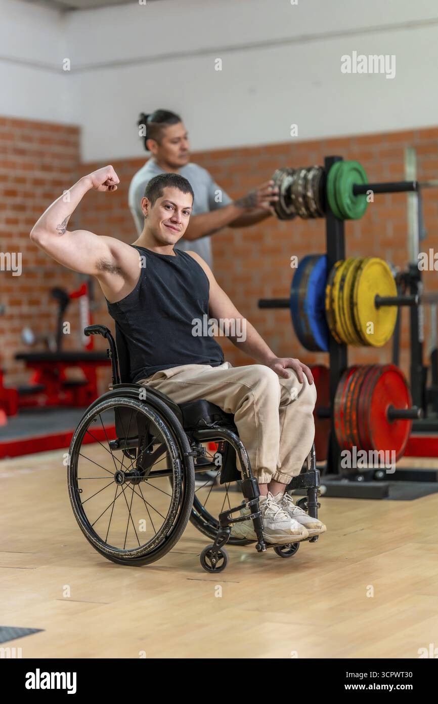 Atleta adattivo su una sedia a rotelle che flessa il bicipite e sorride mentre un allenatore regola le attrezzature per il peso dietro di lui in una palestra luminosa, mostrando forza A. Foto Stock