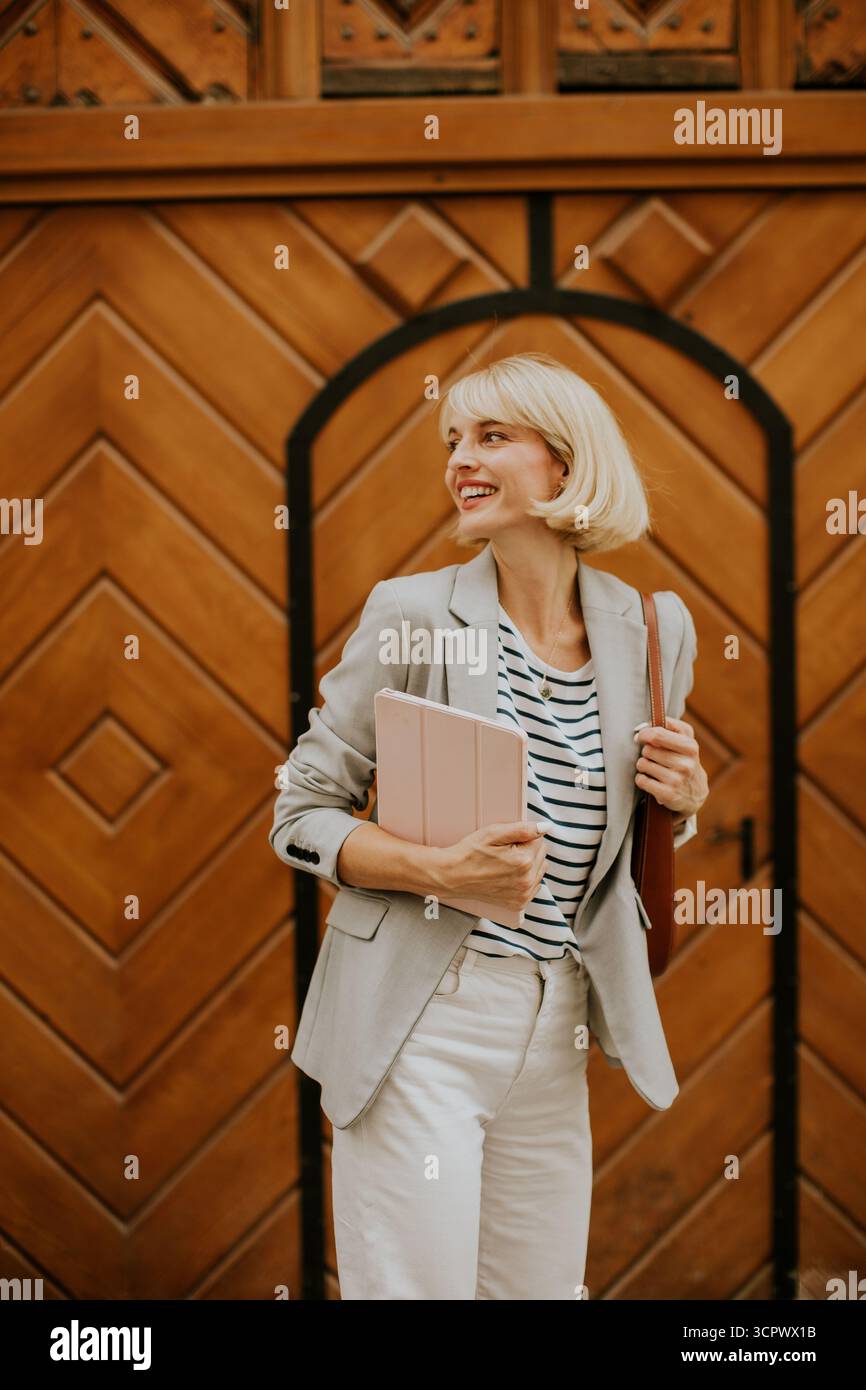 Una donna sicura di sé si trova davanti a una porta di legno, con un blazer e una camicia a righe. Tiene in mano un tablet e una borsa, sorridendo brillantemente in una giornata calda, fa la mostra Foto Stock
