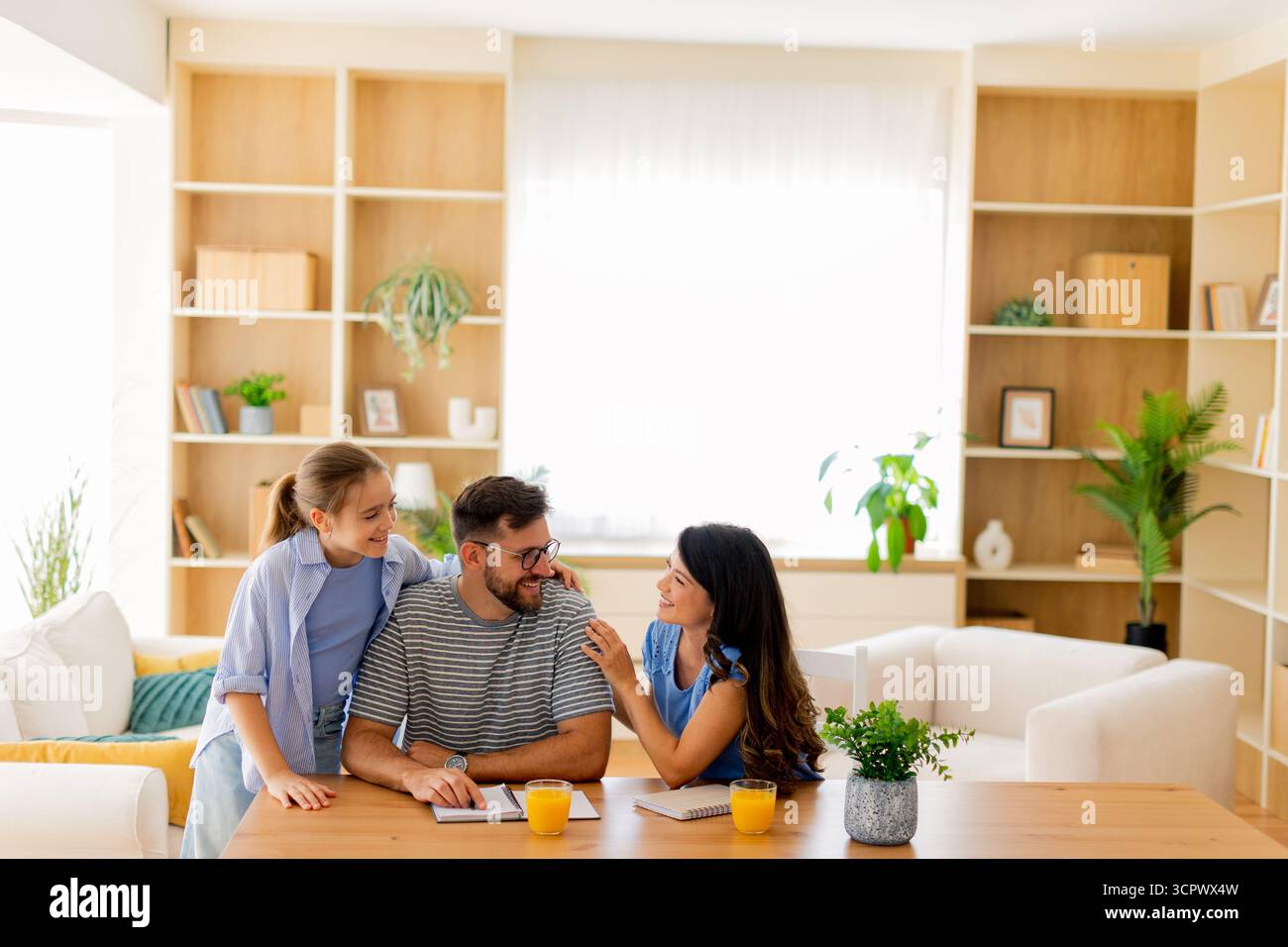 Un momento di gioia si svolge mentre un padre interagisce giocosamente con le sue figlie in uno spazio abitativo illuminato dal sole, circondato da piante e sorrisi. Foto Stock
