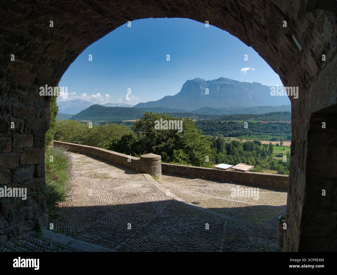 Vista della Valle delle montagne attraverso l'antico Arco di pietra - Una vastissima valle illuminata dal sole e maestose montagne blu incorniciate da un arco di pietra rustico scuro e acciottolato Foto Stock