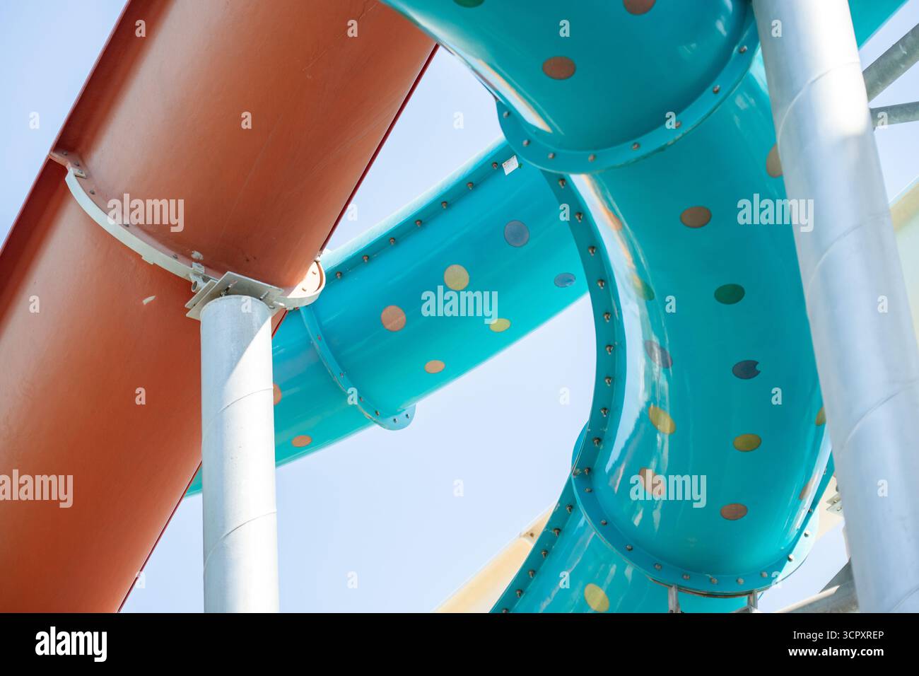 Una foto a basso angolo di scivoli del parco acquatico con tubi arancioni e blu, sostenuti da pali metallici bianchi e grigi, contro un cielo azzurro. Foto Stock