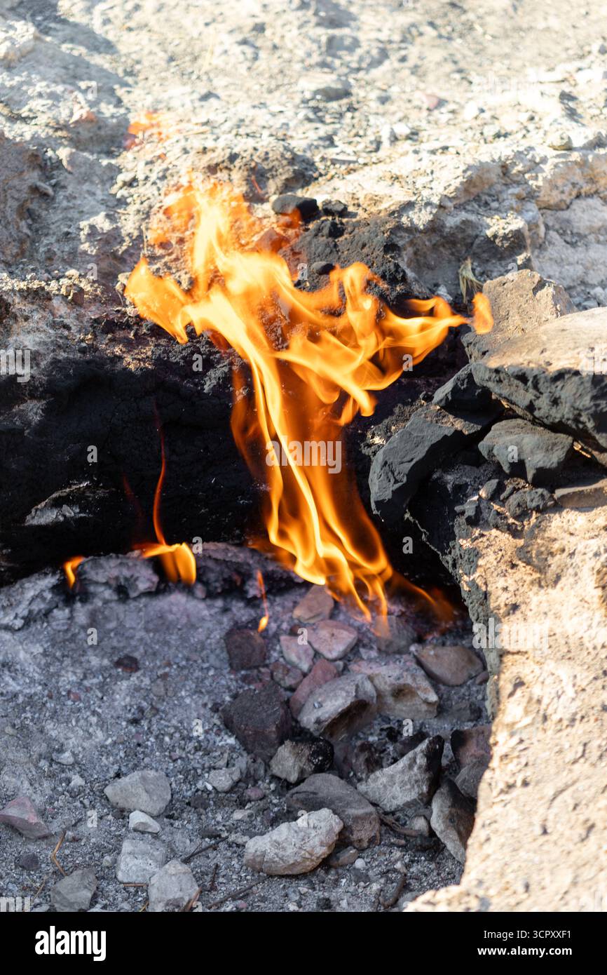 Le fiamme emergono da una fessura rocciosa, contrastando con la cenere circostante, lo sporco e la pietra. La luce solare proietta ombre, evidenziando le trame. Foto Stock
