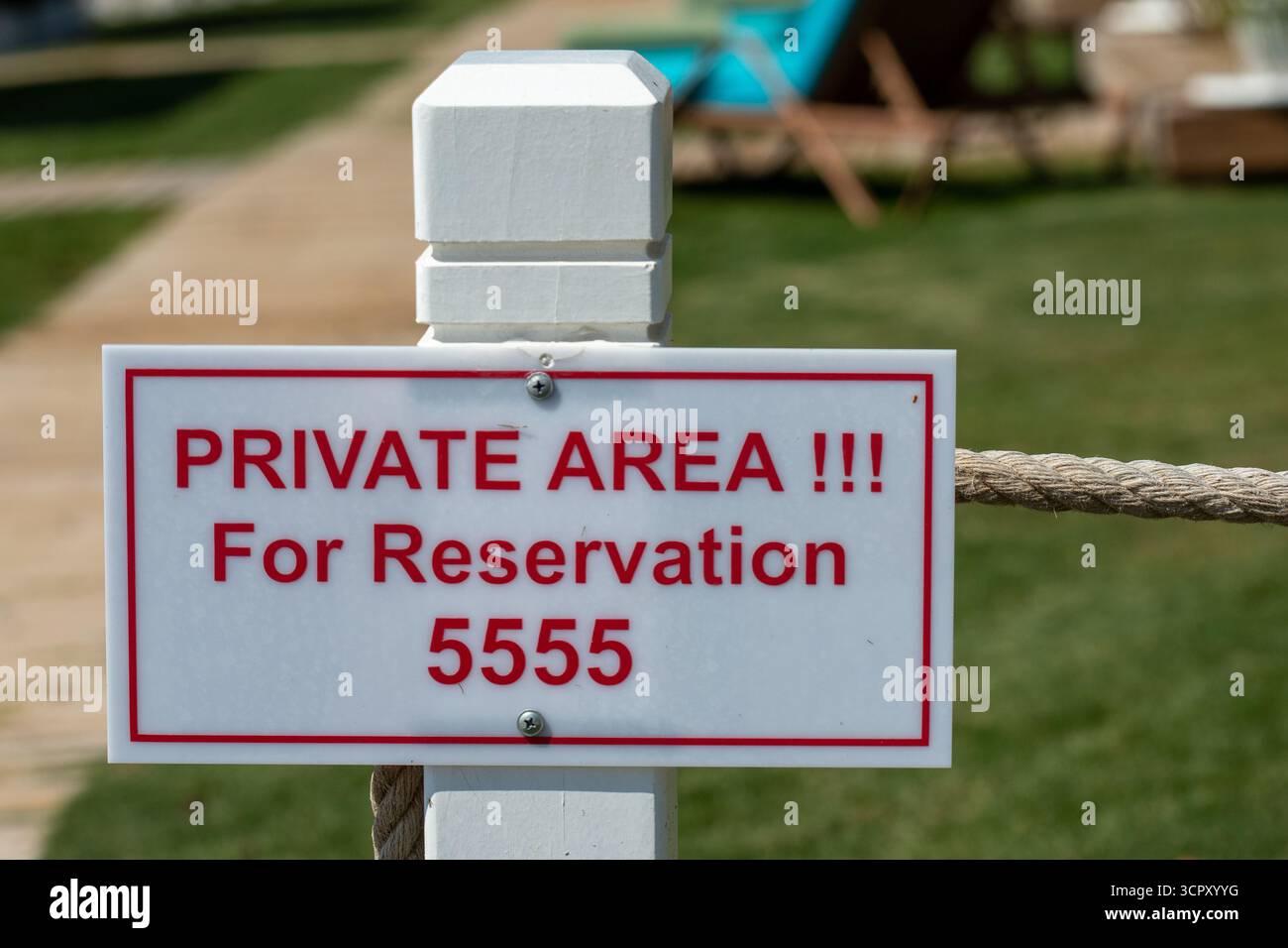 Un cartello con la scritta "area privata!!!" è fissato a un palo bianco con una barriera di corda, adagiato su uno sfondo di erba e un sentiero sfocato. Foto Stock