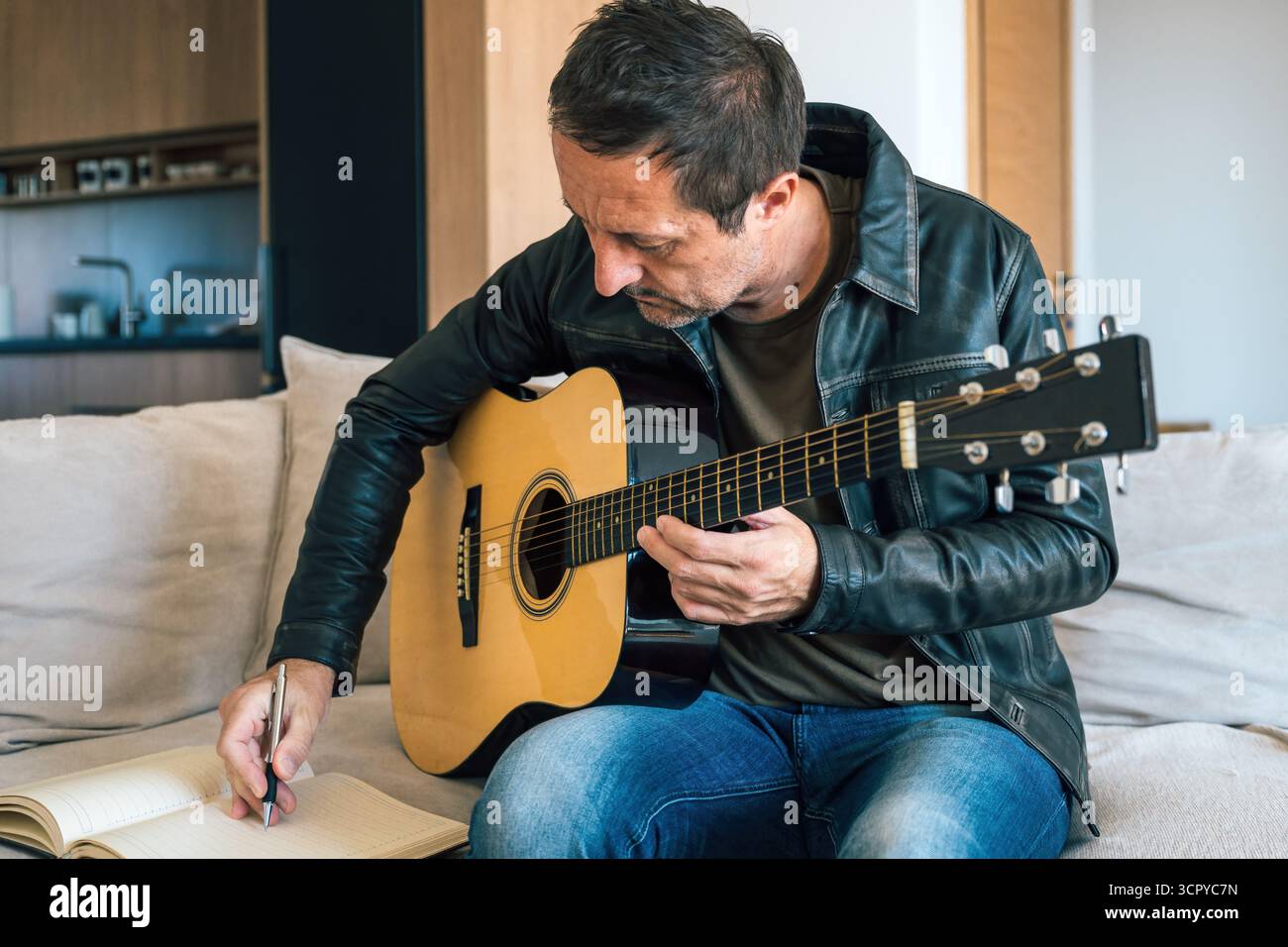 Uomo adulto che compone musica a casa, suona la chitarra e scrive idee su un taccuino, simbolo di espressione artistica, creatività e ispirazione musicale. S Foto Stock