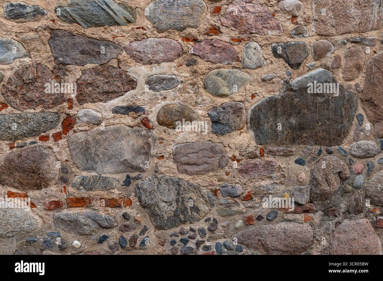 Un muro grosso modo disposto con pietre di varie dimensioni e forme e mattoni rossi. Struttura di pareti in pietra. Parete di ciottoli naturale come sfondo. Foto Stock