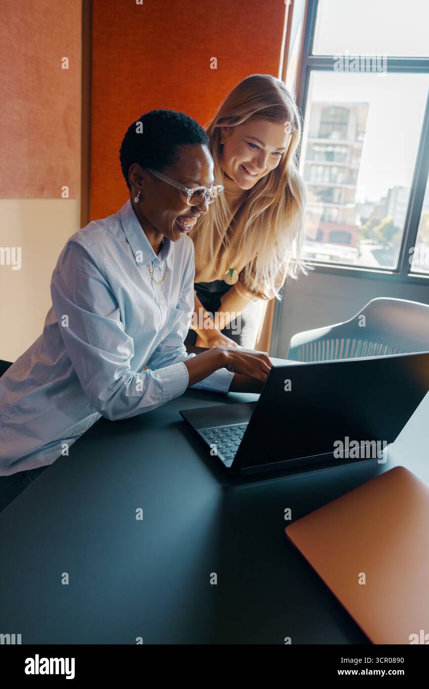 Due donne di diversa provenienza ed età che collaborano a una scrivania in un ufficio. La scena mette in evidenza la cooperazione, l'utilizzo della tecnologia e un lavoro brillante Foto Stock