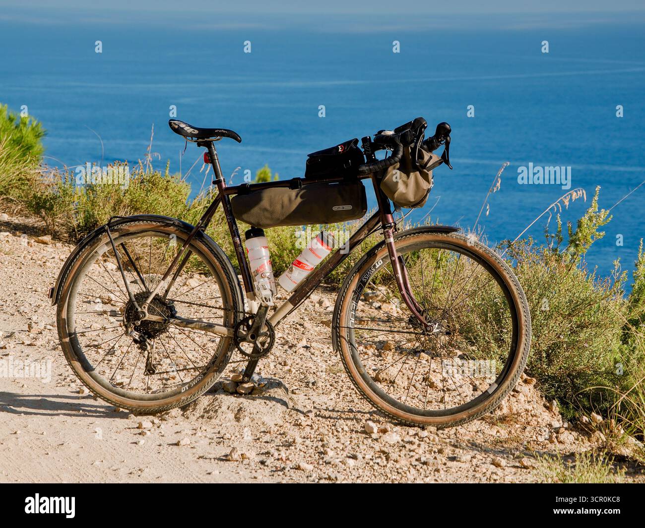 Pedalata ghiaia su un sentiero costiero sterrato sopra il mare blu profondo, viaggio avventuroso in bicicletta alla luce del Mediterraneo estivo. Foto Stock