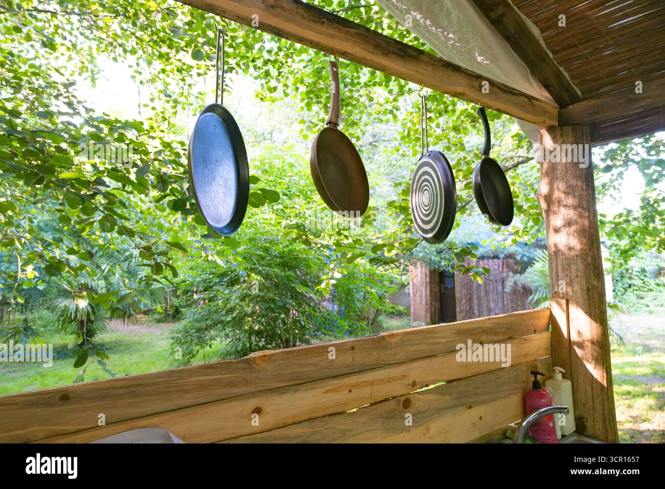Cucina rustica all'aperto con padelle appese e vegetazione lussureggiante. Utensili da cucina appesi all'esterno con sfondo verde Foto Stock