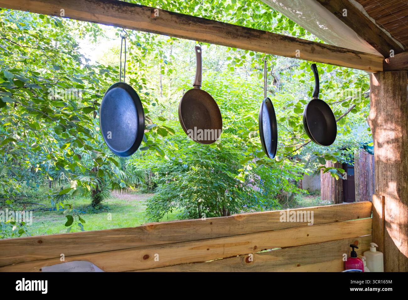Cucina rustica all'aperto con padelle appese e vegetazione lussureggiante. Utensili da cucina appesi all'esterno con sfondo verde Foto Stock