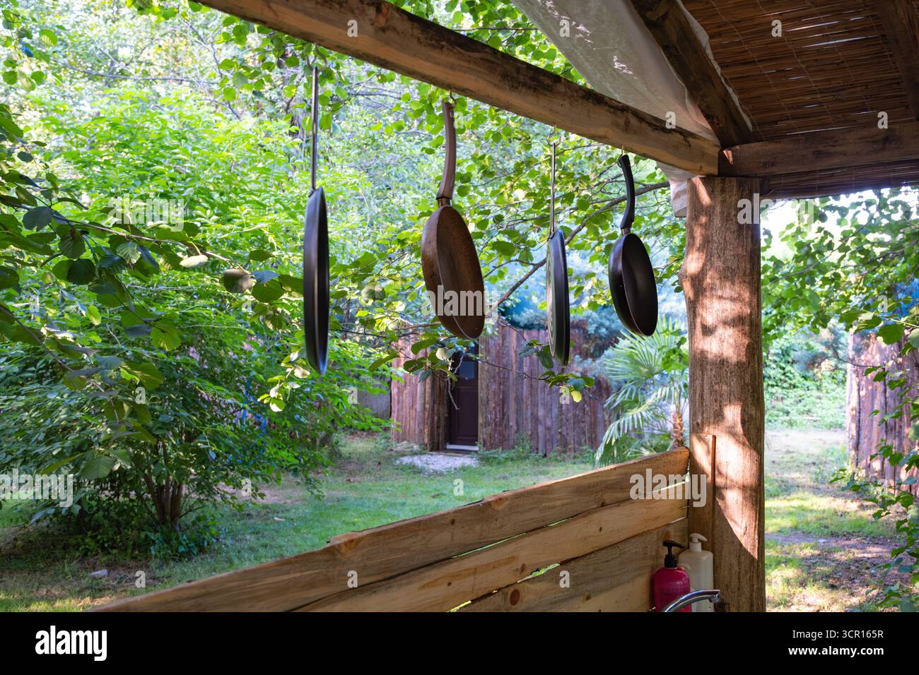 Cucina rustica all'aperto con padelle appese e vegetazione lussureggiante. Utensili da cucina appesi all'esterno con sfondo verde Foto Stock