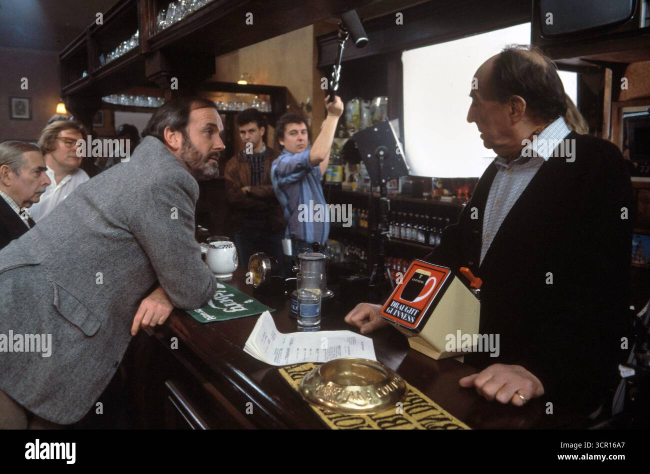 John Cleese esordio nello spot pubblicitario della BBC, Promo Film, conosciuto come la tassa di licenza - Promotional Film, prima messa in onda il 20 gennaio 1986. La pubblicità aveva John Cleese sostenere un pub bar e chiedere 'che cosa ci ha mai dato la BBC? " Ha recitato John Cleese e in questa scena d'apertura Michael Hordern nel ruolo del padrone di casa. E un cast di importanti attori della BBC in un promo per il canone. Londra Inghilterra anni '1985 1980 Regno Unito ( Alan Parker regista (camicia bianca sinistra) questa è la scena di apertura. HOMER SYKES Foto Stock