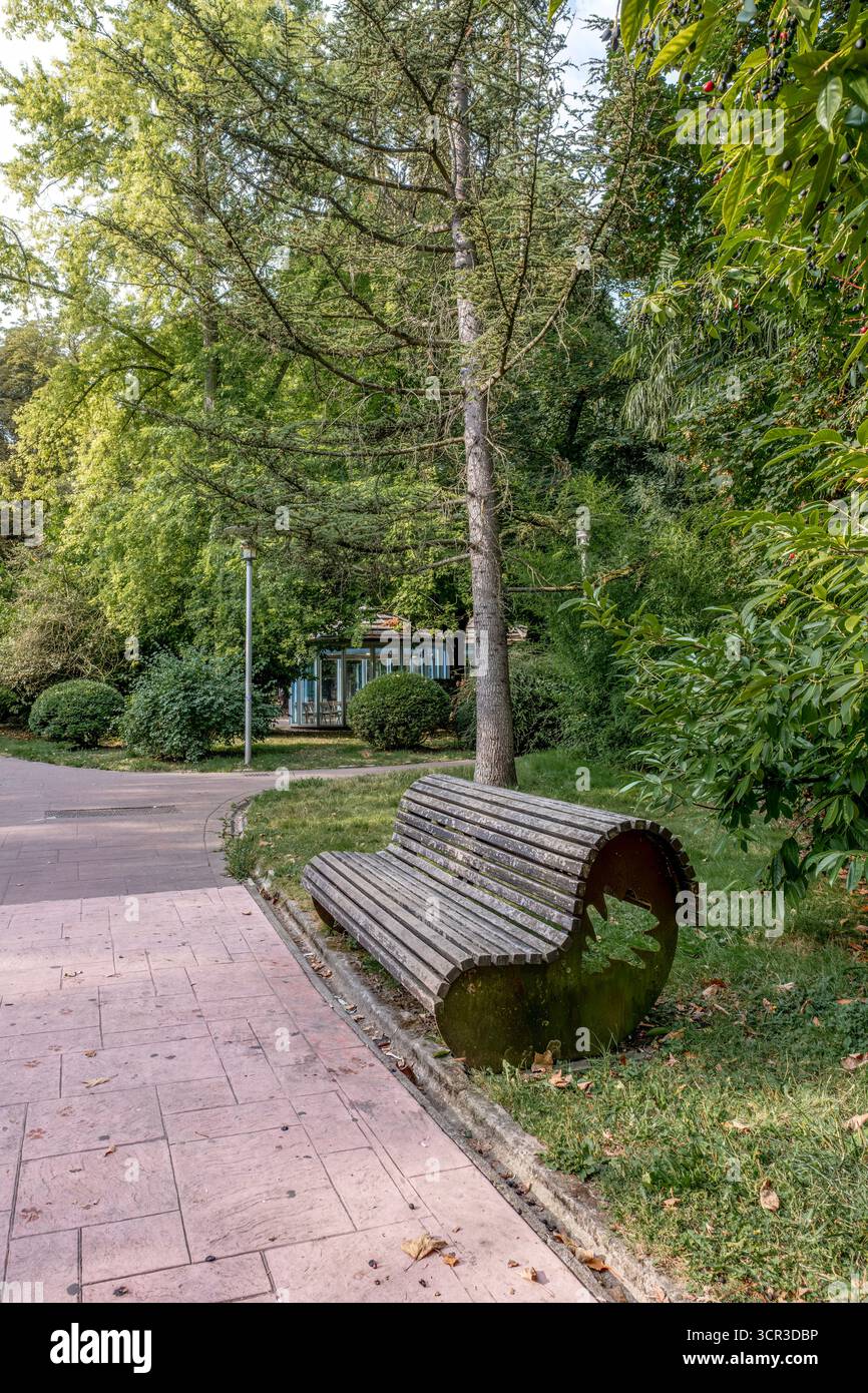 Panca curva in legno su un sentiero lastricato in un parco europeo Shady: Tranquillo spazio verde urbano con lussureggianti folii estivi, alberi e chiosco lontano Foto Stock