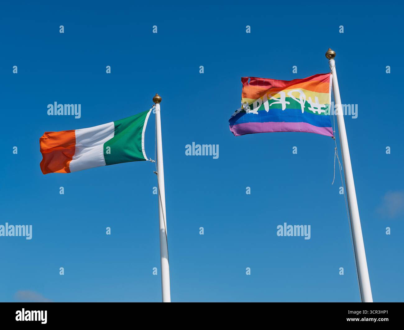 La bandiera nazionale irlandese e la bandiera dell'arcobaleno si abbinano a pali bianchi. Il cielo è blu chiaro. Situato a West Cork, Irlanda. Foto Stock