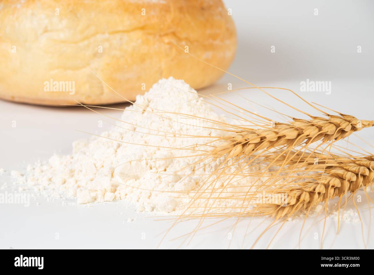 Mucchio di farina accanto ai gambi di grano essiccato con un formaggio rotondo sullo sfondo, evidenziando gli ingredienti essenziali della cucina. Foto Stock