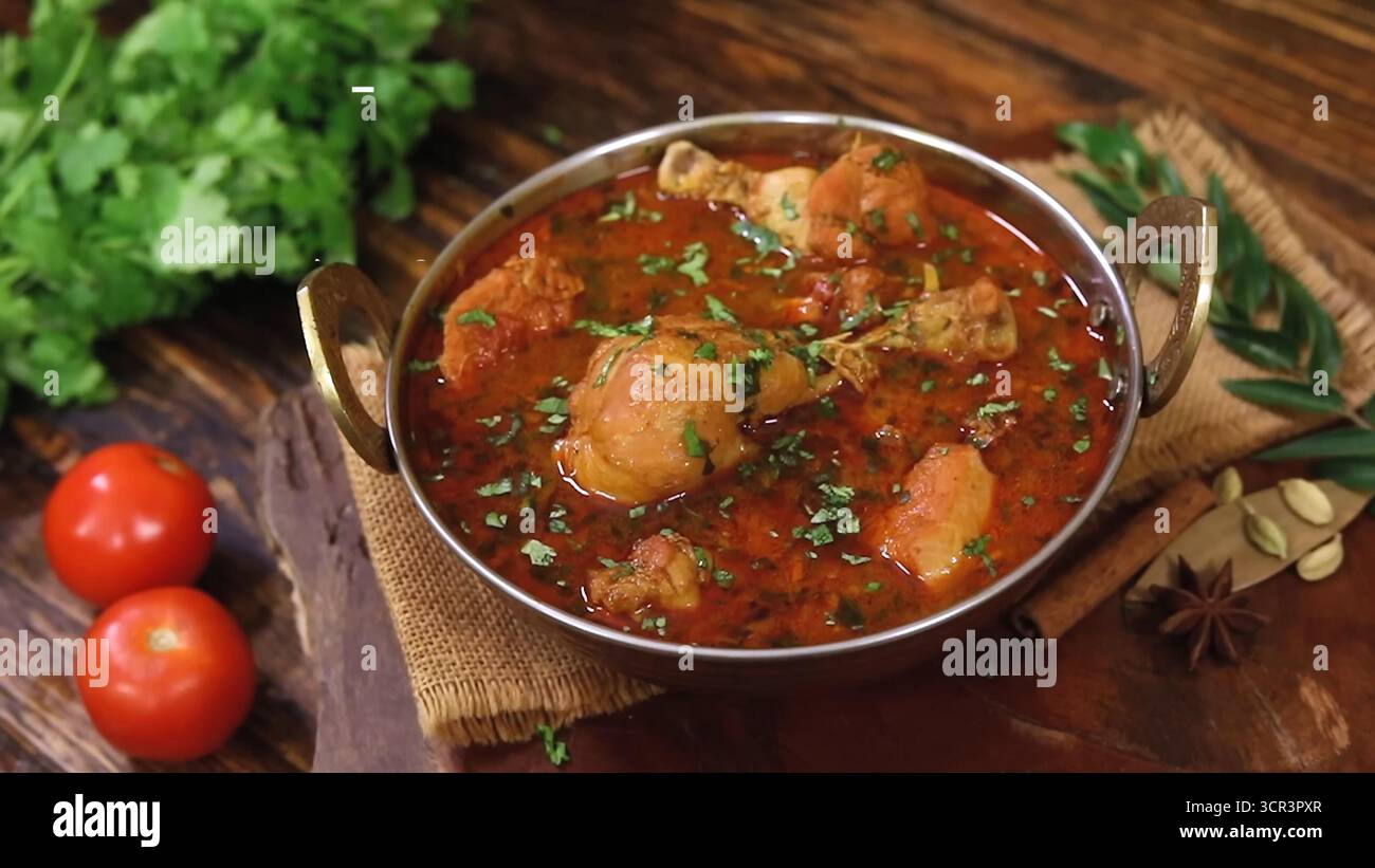 Pollo al curry servito in una teglia al curry, guarnito con foglie di coriandolo fresco. Un tavolo marrone scuro con verdure che ne esaltano il fascino Foto Stock