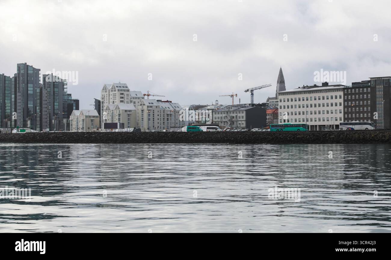 Una tranquilla zona portuale lungo un lungomare mostra un mix di edifici moderni e tradizionali, un gabbiano solitario che vola sopra l'acqua e gru distanti Foto Stock Una tranquilla zona portuale lungo un lungomare mostra un mix di edifici moderni e tradizionali, un gabbiano solitario che vola sopra l'acqua e gru distanti Foto Stock