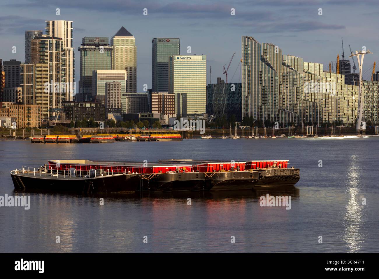 Skyline della città illuminato dal sole con alti edifici che si riflettono sulle calme acque del fiume sotto un cielo azzurro. Canary Wharf, Londra, Regno Unito Foto Stock