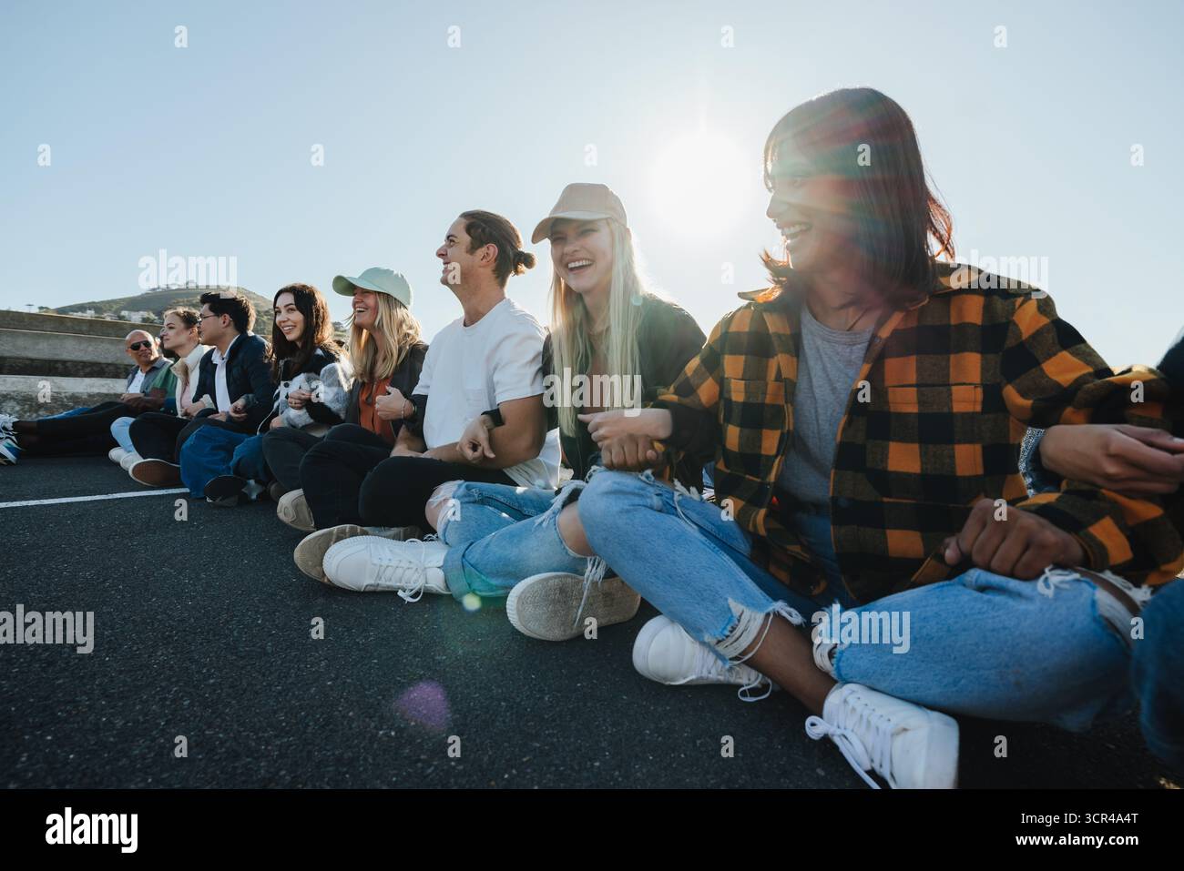 Un gruppo variegato di giovani adulti che partecipano a una pacifica protesta all'aperto, tenendosi per mano mentre sono seduti insieme su una strada. L'atmosfera lo trasmette Foto Stock Un gruppo variegato di giovani adulti che partecipano a una pacifica protesta all'aperto, tenendosi per mano mentre sono seduti insieme su una strada. L'atmosfera lo trasmette Foto Stock