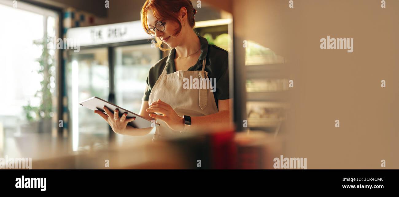 L'imprenditore che indossa un grembiule utilizza un tablet per gestire l'inventario e gli ordini dei clienti in una gastronomia moderna. Concetto di proprietà di piccole imprese, ristorante Foto Stock