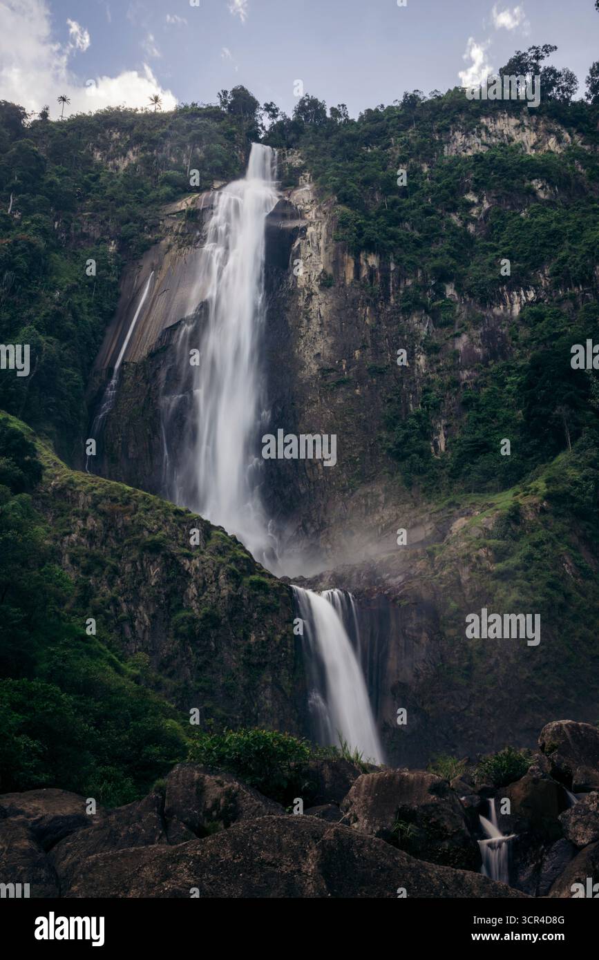 La maestosa cascata di Air Terjun Nirwana Wartefall scende giù da una lussureggiante montagna verde sotto un cielo blu brillante. Toba, Sumatra settentrionale, Indonesia Foto Stock