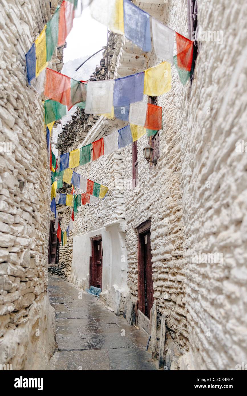 Vicolo stretto in pietra adornato da colorate bandiere di preghiera tra edifici rustici intonacati. Marpha, Annapurna, distretto di Mustang della provincia di Gandaki, Nepal Foto Stock